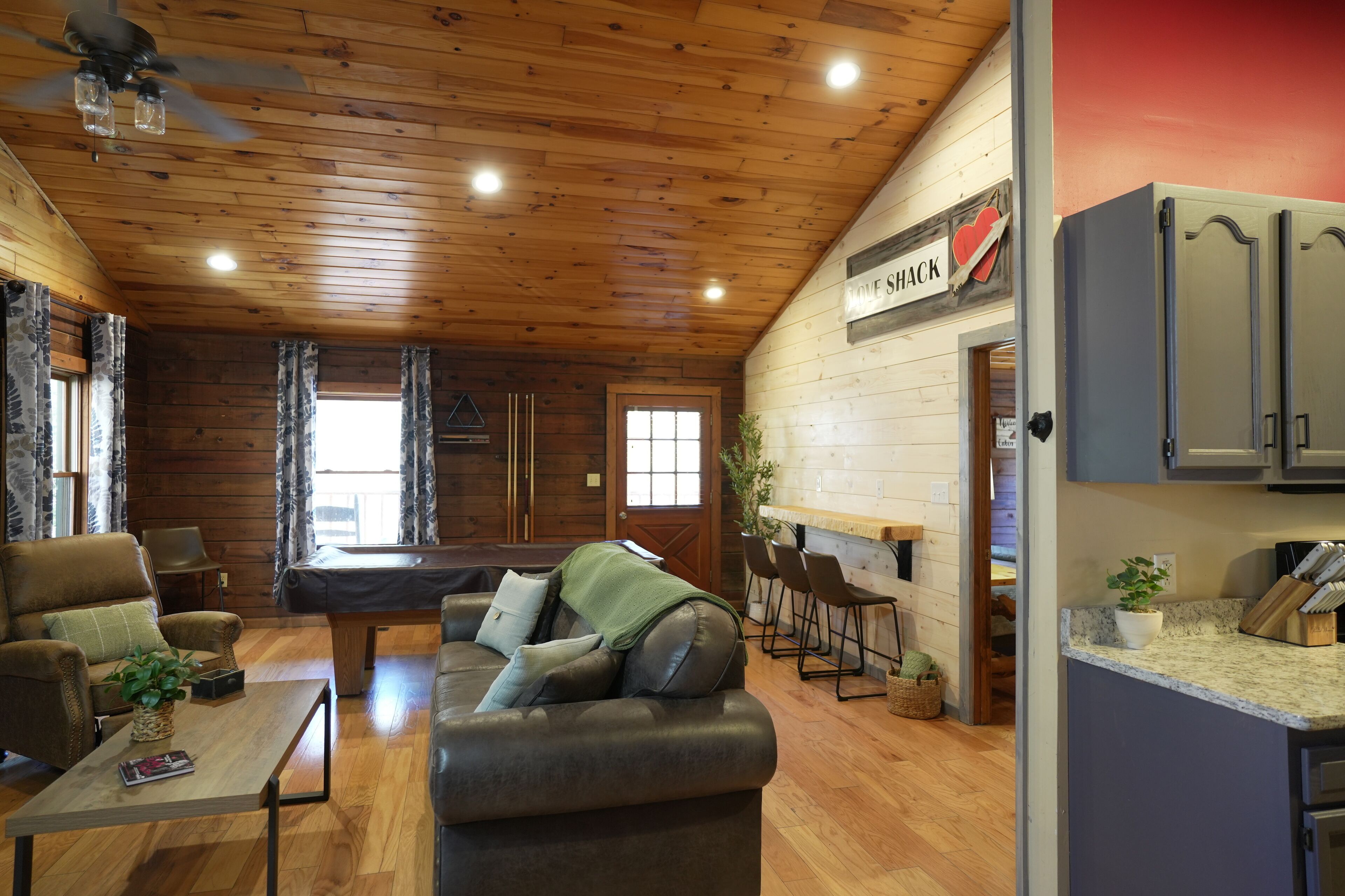 A cozy cabin interior with a wooden ceiling, a billiards table, a sofa, and a kitchen area with bar stools.