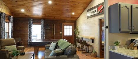 A cozy cabin interior with a wooden ceiling, a billiards table, a sofa, and a kitchen area with bar stools.