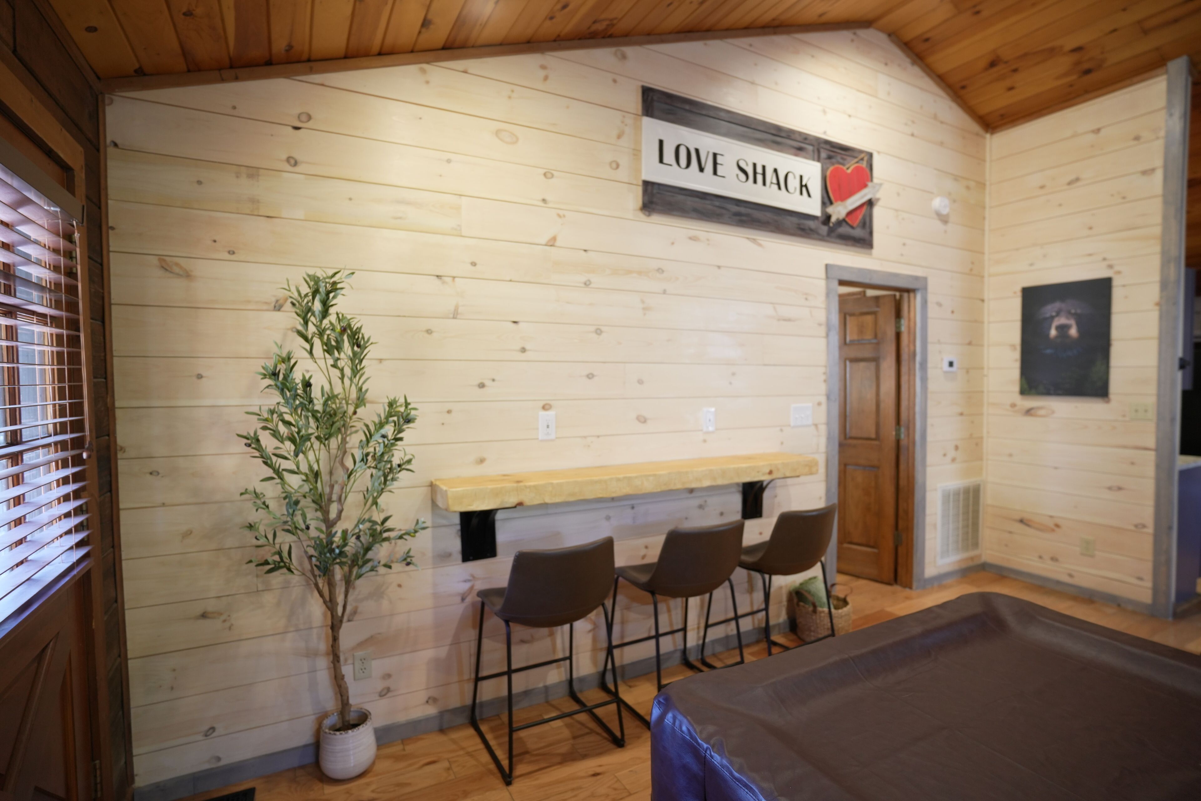 Ett mysigt rum med träväggar, en bardisk med barstolar och en skylt med texten ”LOVE SHACK”.