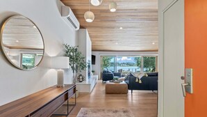 Un salon moderne avec un plafond en bois, un grand miroir rond, une console en bois, une grande fenêtre donnant sur l’extérieur et un coin salon comprenant un canapé et un pouf.