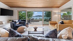Un vaste salon doté d’un plafond en bois, d’un grand téléviseur fixé au mur et offrant une vue sur un lac grâce à de grandes portes-fenêtres coulissantes.