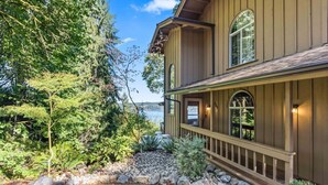 A two-story house with a stone pathway leading to the front door, surrounded by lush greenery and a view of a lake. - Eagle`s Lookout Lodge w/ Hot Tub (Gig Harbor)