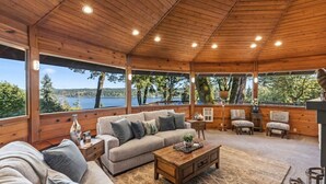 A cozy cabin with a wooden interior, large windows offering a view of a lake, a sofa, a coffee table, and two chairs. - Eagle`s Lookout Lodge w/ Hot Tub (Gig Harbor)