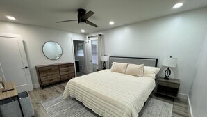 Un dormitorio con una cama grande, un ventilador de techo, una mesita de noche con lámpara, un aparador y un espejo en la pared.