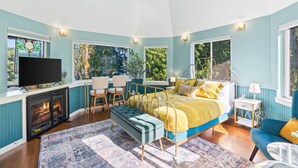 A bedroom with a bed, bedside table, lamp, TV, fireplace, and windows.