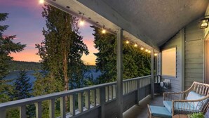 A balcony with a view of a lake and forest, featuring a wicker chair and a small table with a plant.