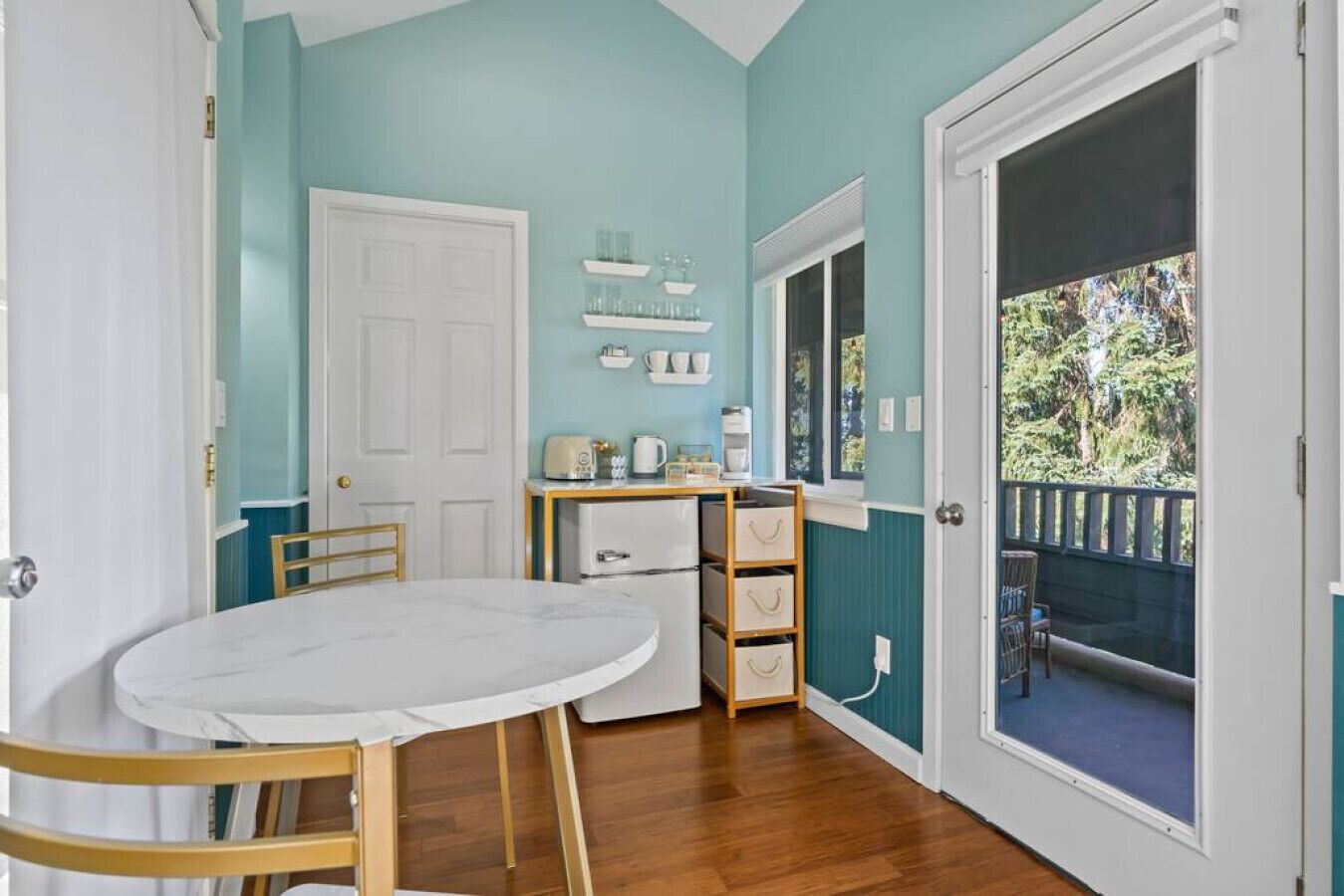 A small dining area with a round table and chairs, a white door, a window with a view of greenery, and a shelf with kitchen items.