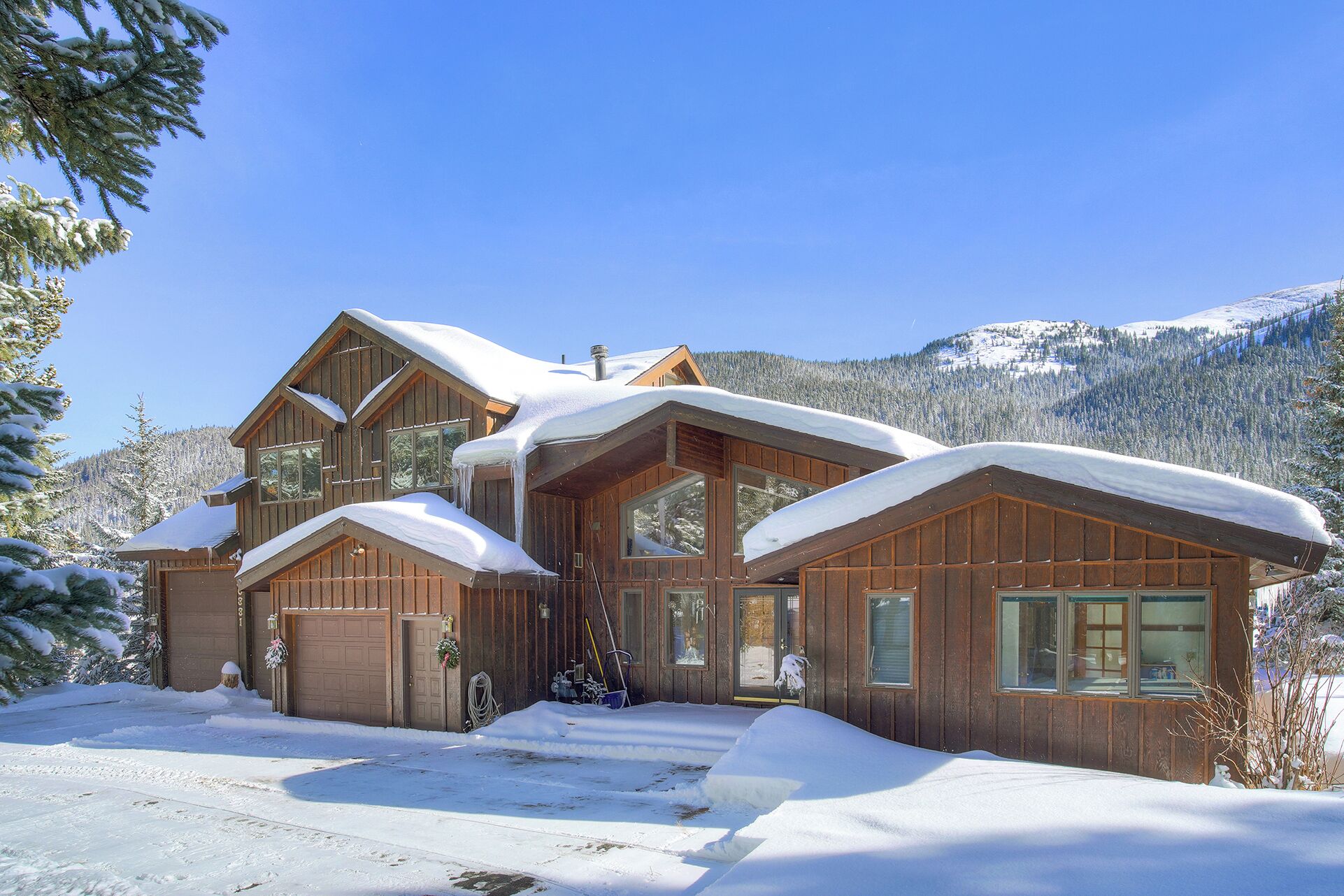 A wooden cabin with a garage, large windows, and a snowy mountain backdrop.