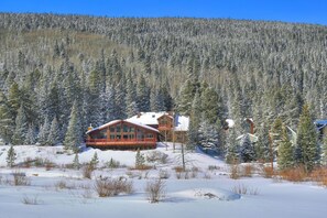 A log cabin with a large deck, surrounded by a forested area with snow-covered trees.
