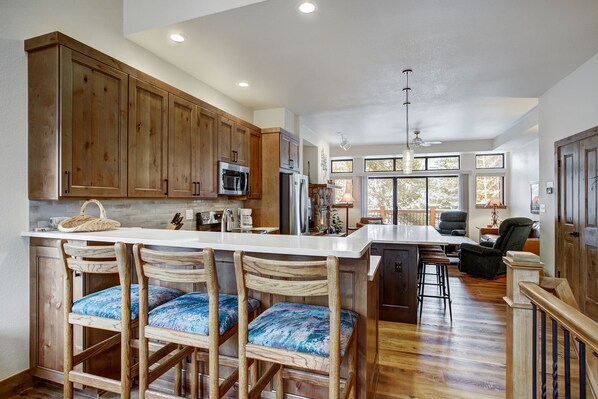 A spacious kitchen with wooden cabinets, a central island with bar stools, a stone fireplace, and large windows offering views of trees. - Village Point 308 (Breckenridge)