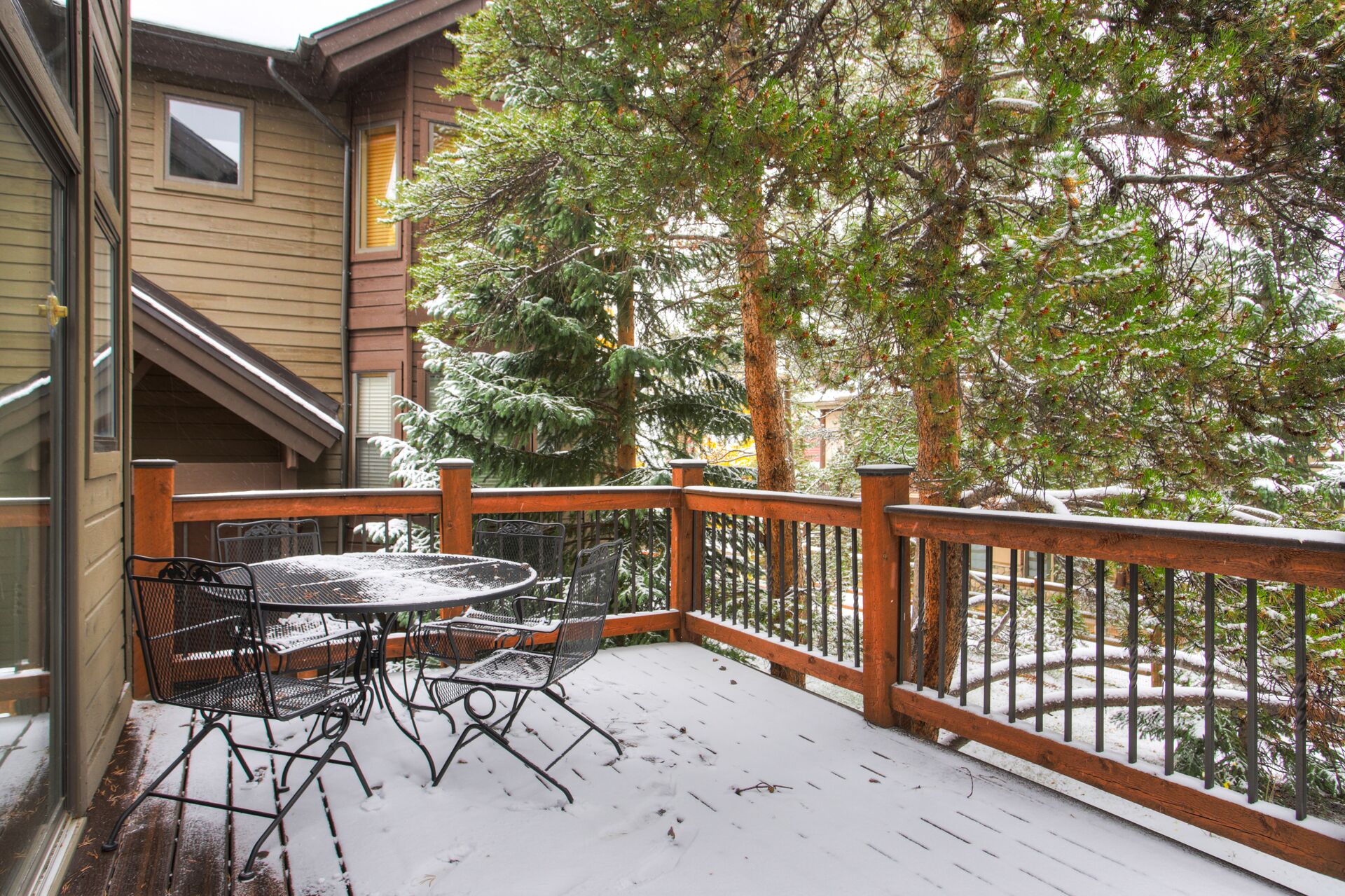A wooden deck with metal chairs and a table, surrounded by snow-covered trees.