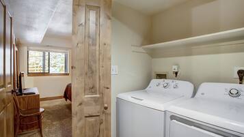 Un lavadero con dos lavadoras blancas, una puerta de madera, una silla y una ventana con vistas a árboles.