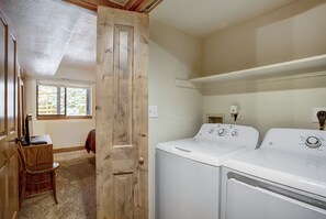A laundry room with two white washing machines, a wooden door, a chair, and a window with a view of trees. - Village Point 308 (Breckenridge)