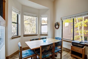 A dining area with a wooden table and blue upholstered chairs, large windows with views of trees, and a printer on a wooden stand. - Village Point 308 (Breckenridge)
