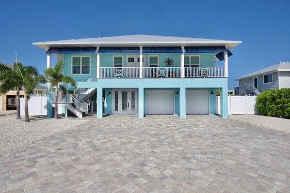 A two-story house with a blue exterior, a balcony with white railings, and a driveway with patterned paving.