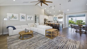 A spacious living room with a sectional sofa, a coffee table, and a ceiling fan. There is a kitchen area with a dining table and chairs visible through an open door. - Crain's Perch will Wow you! (Fort Myers Beach)