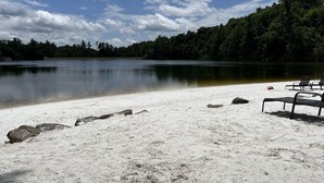 Una playa de arena blanca, un lago tranquilo y una zona boscosa al fondo. Hay sillones de playa sobre la arena.
