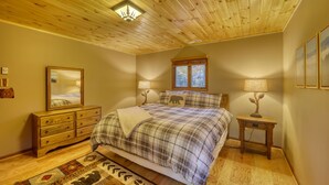 Un dormitorio con techo de madera, una cama con cubrecama a cuadros, una mesita de noche con una lámpara, un aparador y un espejo.