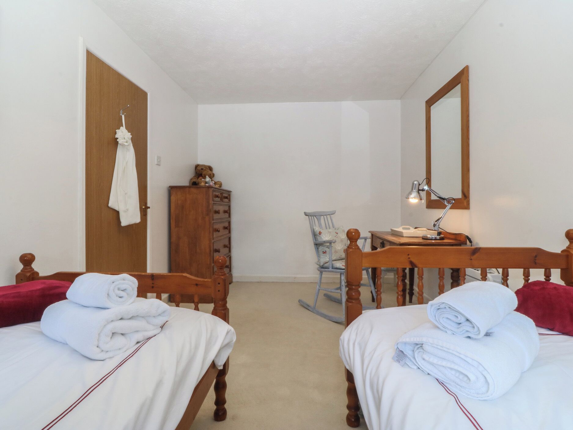 A bedroom with two beds, a wooden dresser, a rocking chair, a desk with a lamp, and a mirror on the wall.