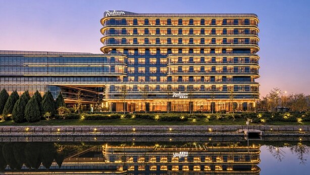 A multi-story Radisson hotel with a modern glass facade, a reflection in the water, and a landscaped front area. - Radisson Hotel, Beijing Daxing Airport     (Daxing)