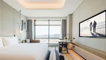 A modern hotel room with a large bed, a desk, a chair, a TV screen, and a view of the city through large windows.