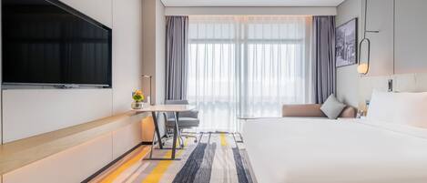 A modern hotel room with a large bed, a desk with a chair, a flat-screen TV mounted on the wall, and a large window with curtains.