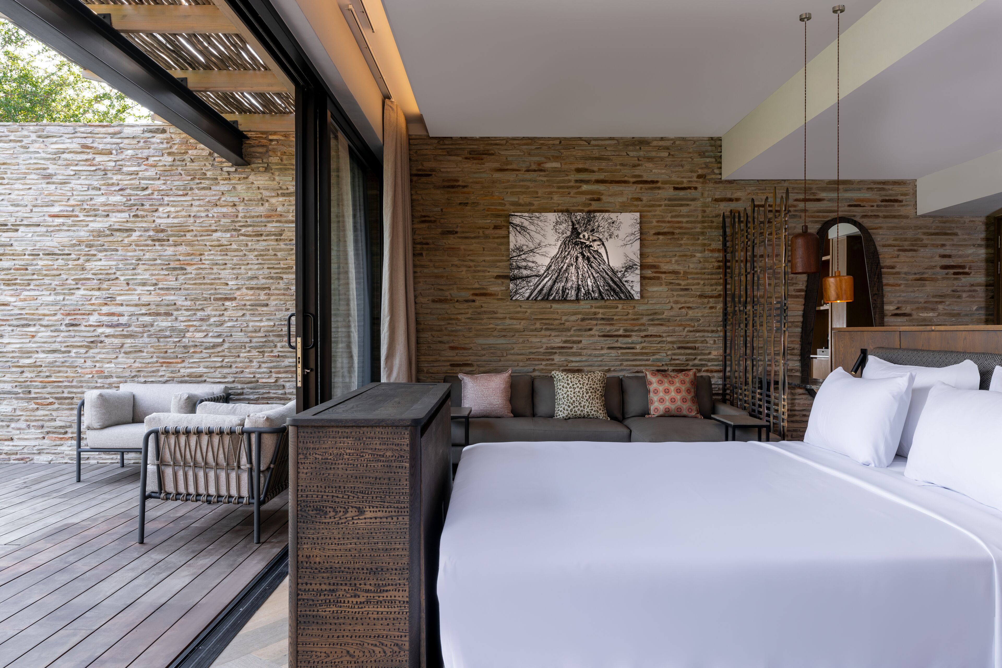 Une chambre d’hôtel moderne avec un grand lit, un canapé et une petite table. On y trouve un mur en pierre et une terrasse en bois à l’extérieur.
