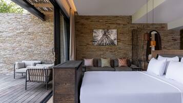 Une chambre d’hôtel moderne avec un grand lit, un canapé et une petite table. On y trouve un mur en pierre et une terrasse en bois à l’extérieur.