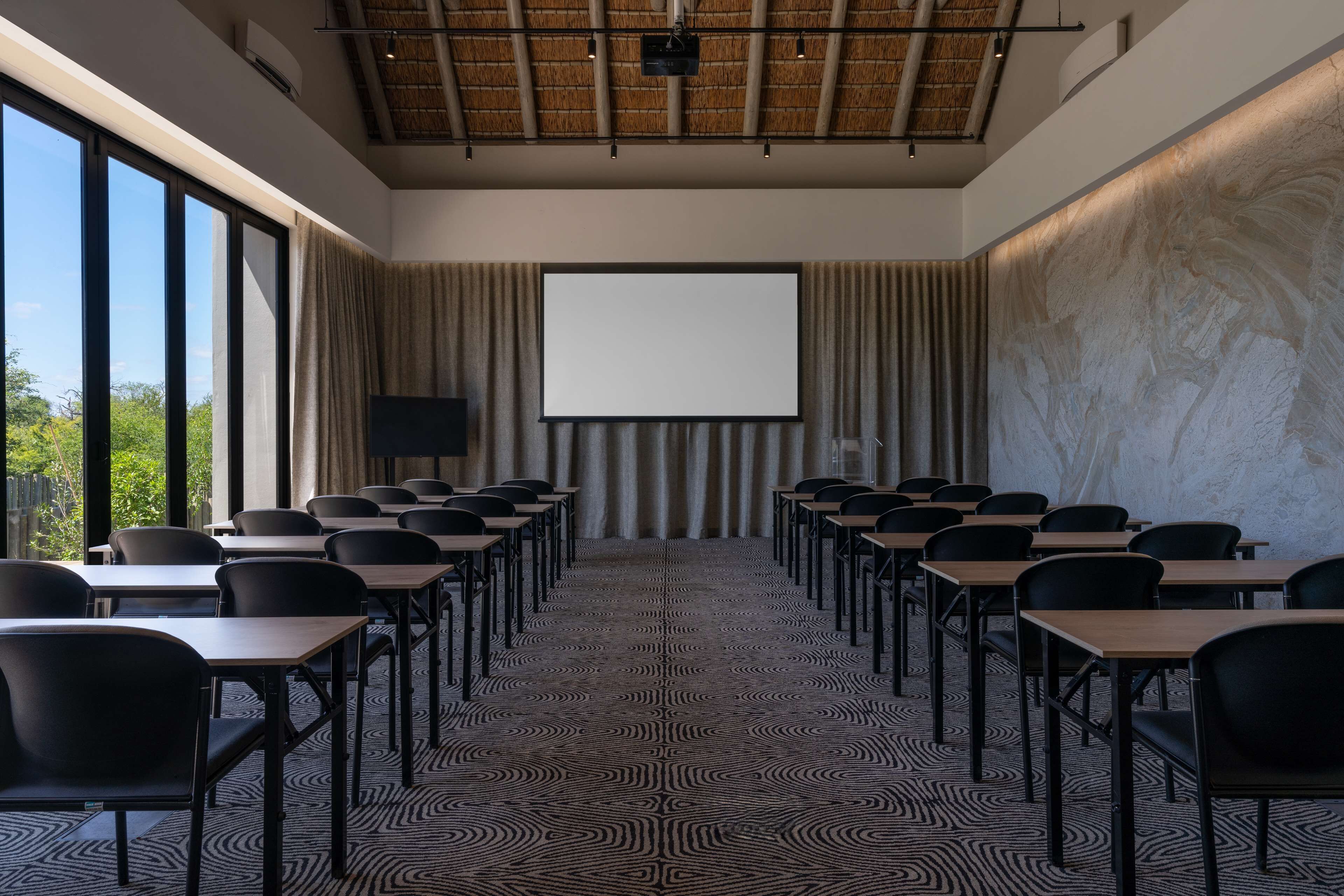 Uma sala de conferências com uma tela grande, fileiras de cadeiras e um tapete estampado.