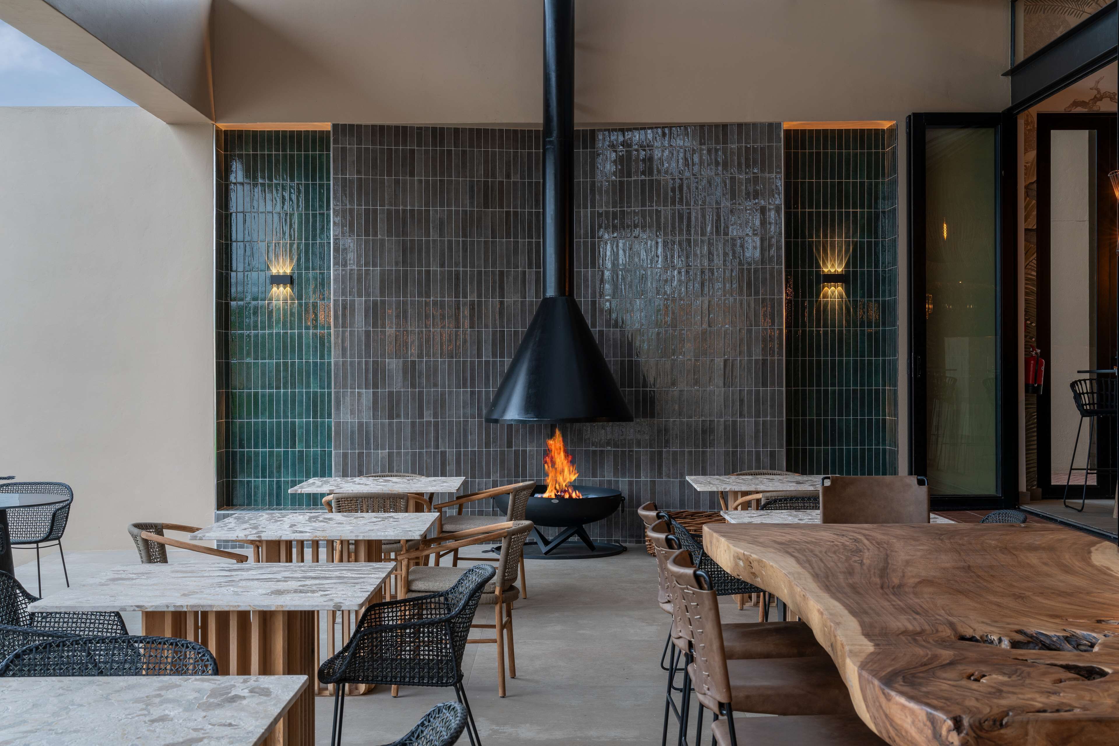 A modern outdoor dining area with a central fire feature, surrounded by wooden tables and chairs.