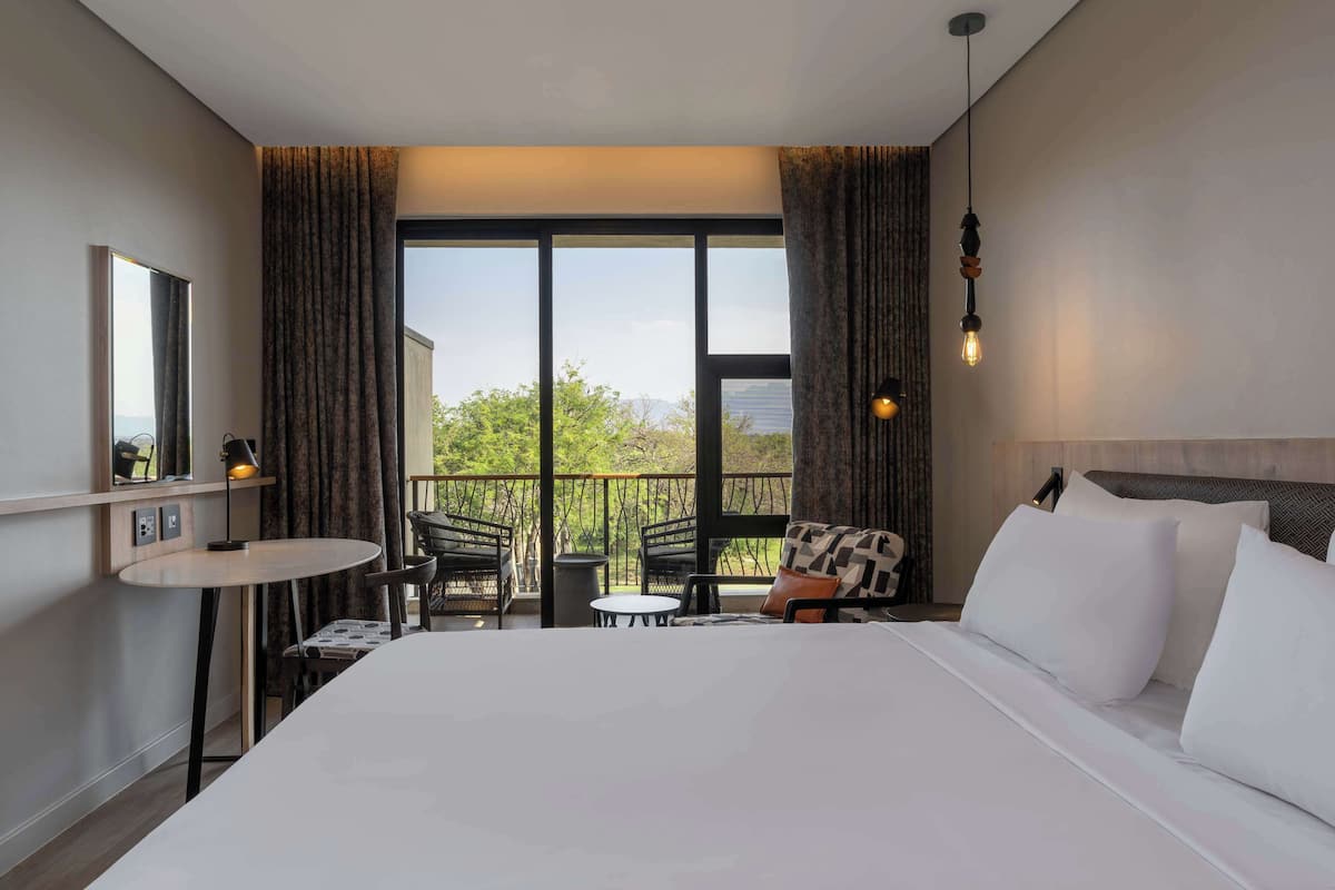 A hotel room with a large bed, a balcony with seating, and a view of trees and buildings.