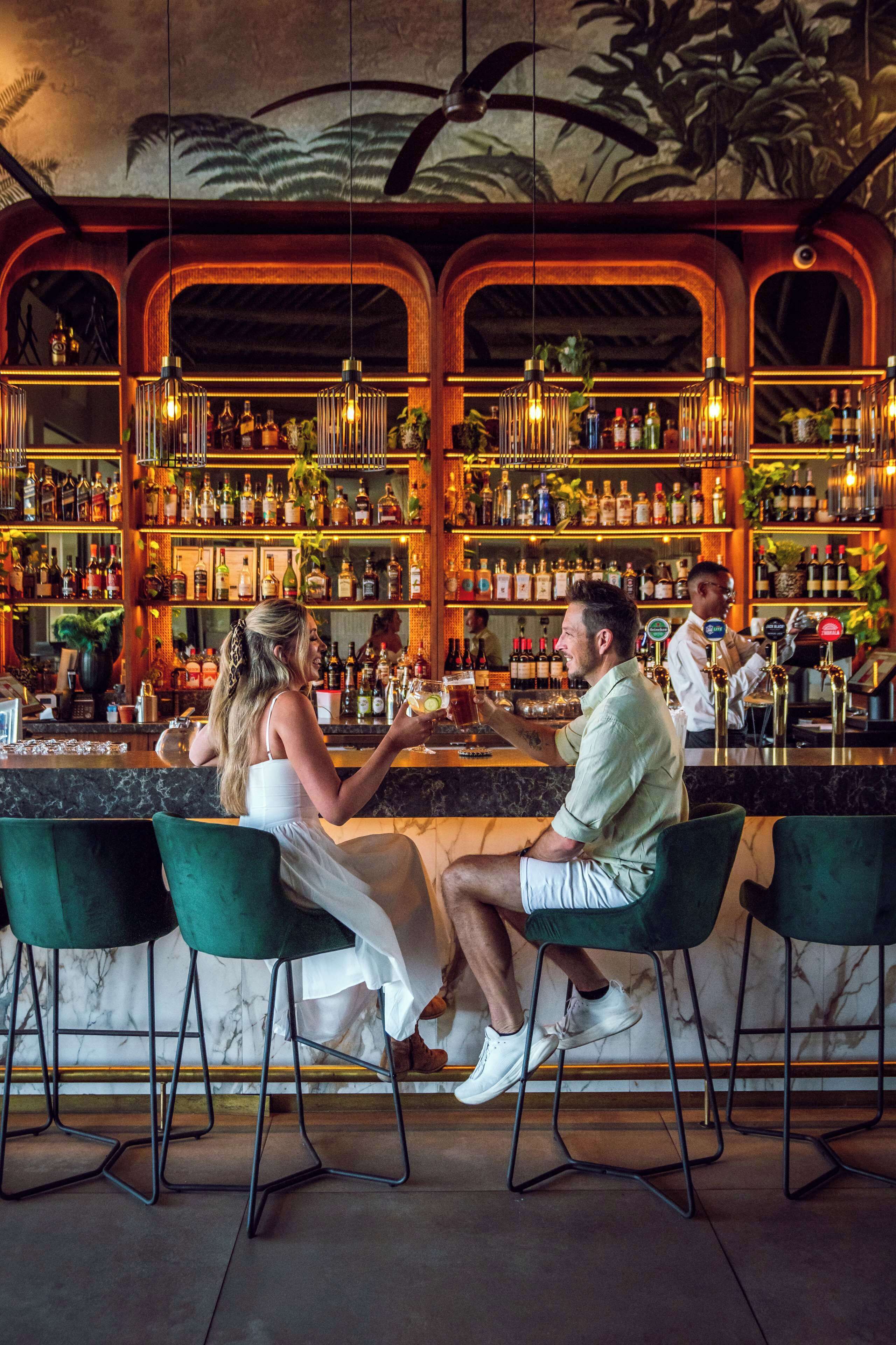 A bar with a high bar counter, multiple shelves of bottles, and two patrons seated at the bar.