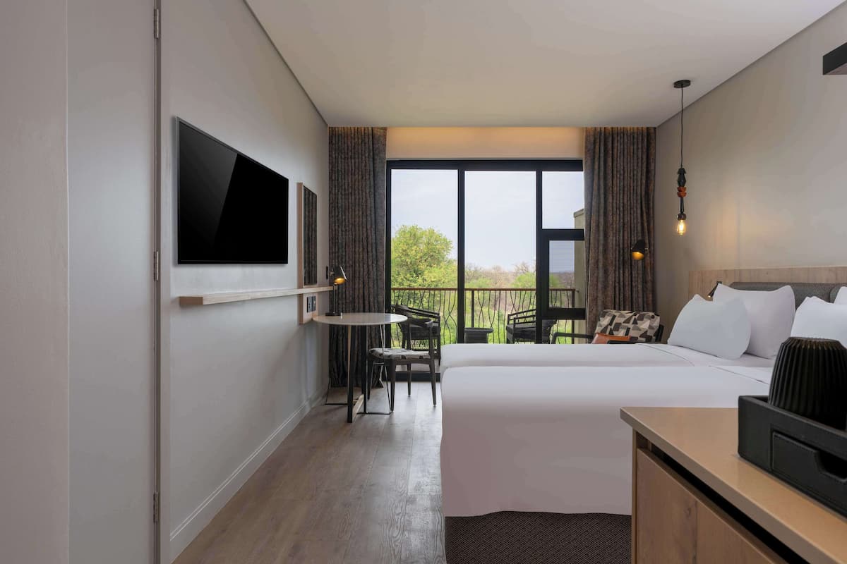 A hotel room with a large bed, a flat-screen TV mounted on the wall, a small round table with chairs, and a balcony with a view of greenery.