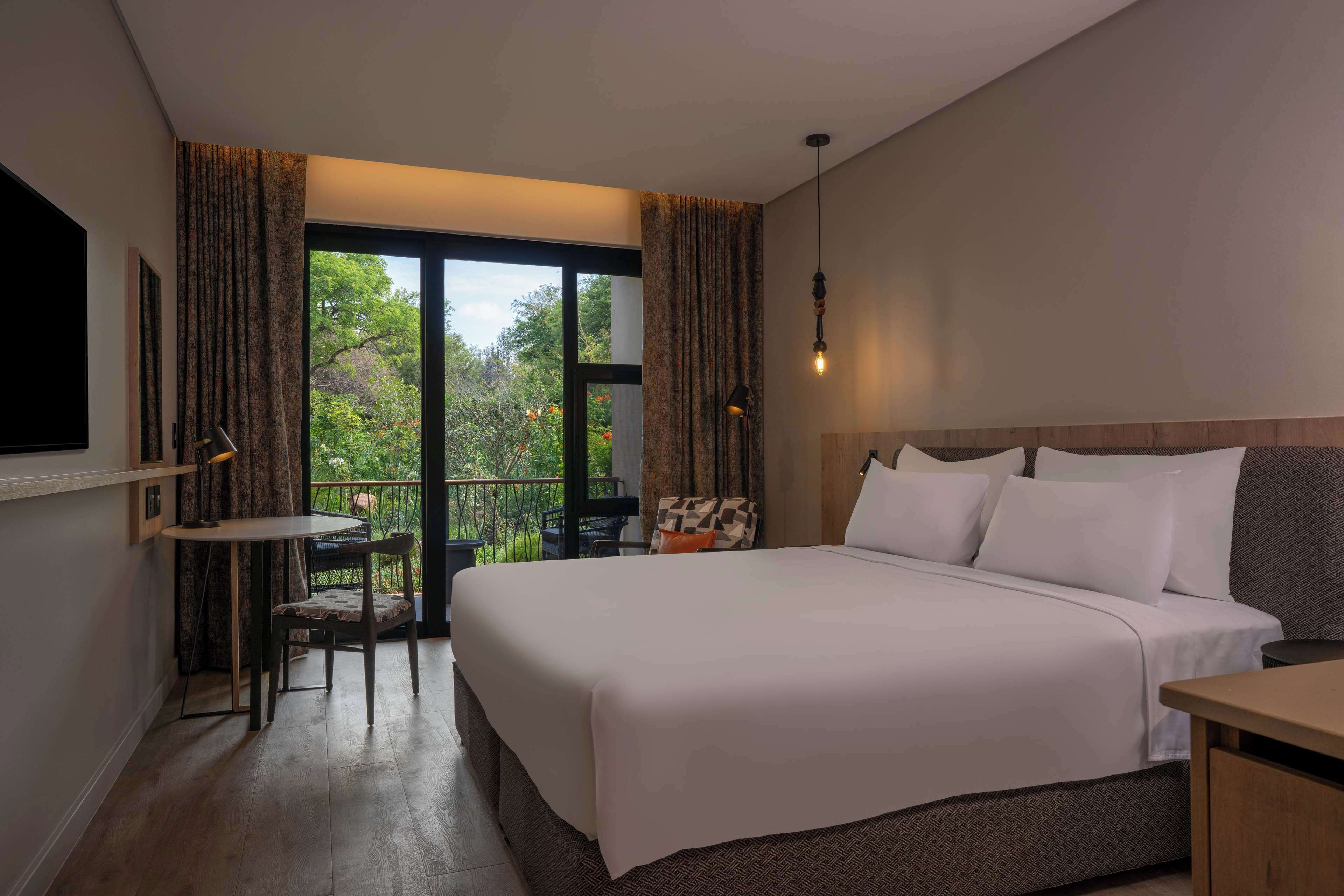 Ein Hotelzimmer mit einem großen Bett, einem kleinen Tisch und einem Stuhl. Es gibt einen Balkon mit Blick auf Bäume und einem Geländer.