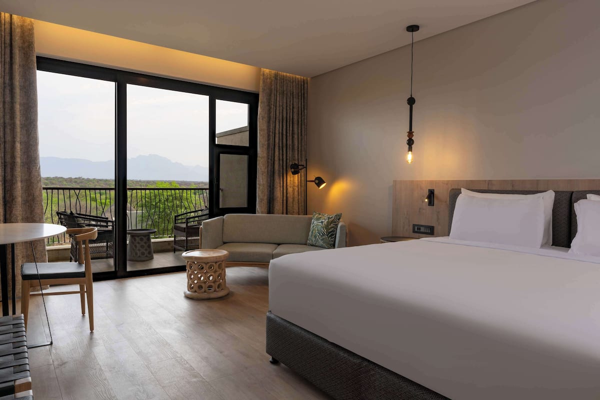 A hotel room with a large bed, a sofa, a dining table, and a balcony with a view of the mountains.