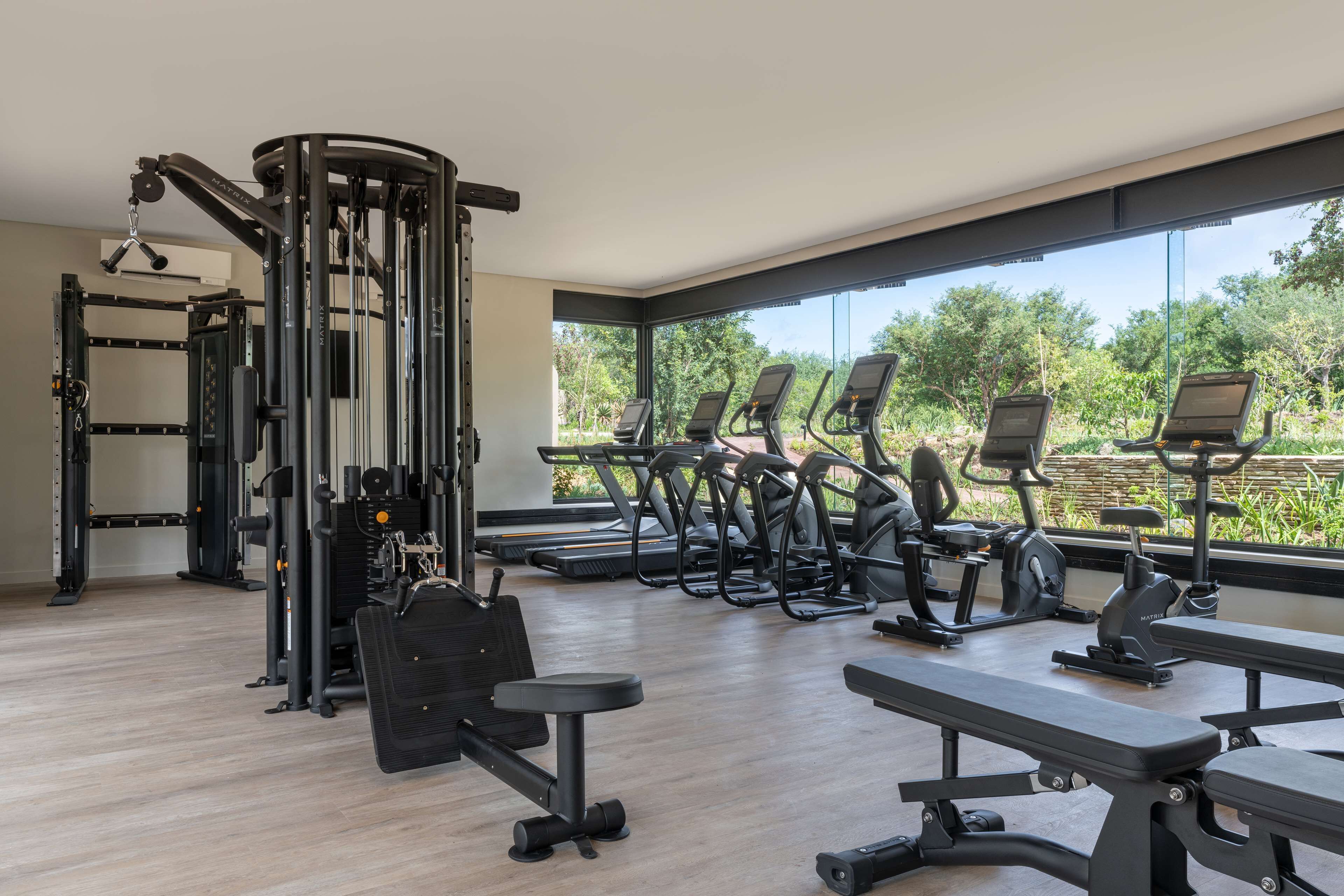 A modern gym with various exercise equipment, large windows, and a view of greenery.