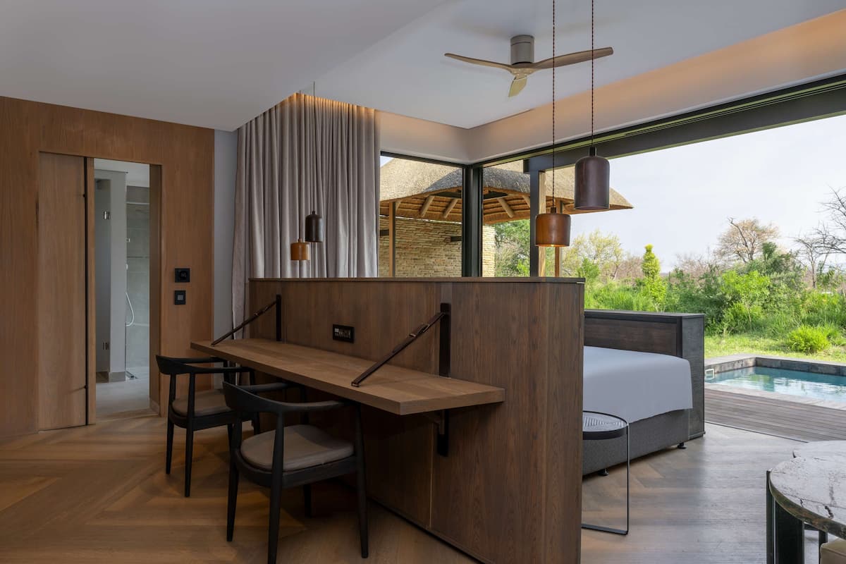 A modern room with a wooden desk, chairs, a bed, and a view of a pool and greenery.