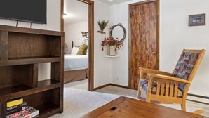 A room with a wooden bookshelf, a wooden coffee table, a patterned armchair, a bed, a TV, and a wooden door. - Outpost: Vine Street 3 - Close to Snow King (Jackson)