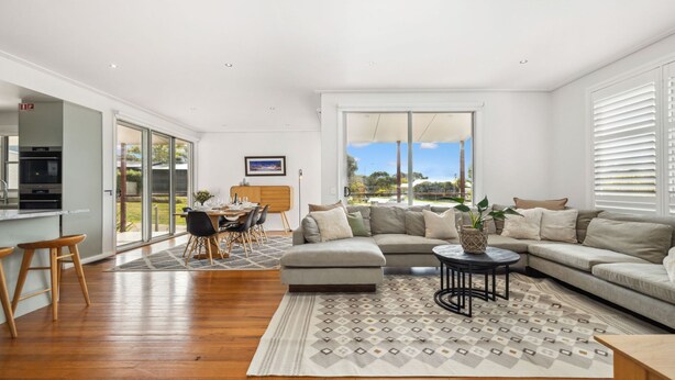 A spacious living area with a large sofa, a dining table, and a kitchen area with a built-in oven. - Alpine Snow Angels Retreat (Jindabyne)