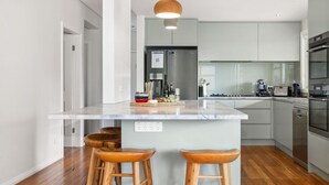 A modern kitchen with a marble island, wooden stools, stainless steel appliances, and a view into the dining area. - Alpine Snow Angels Retreat (Jindabyne)