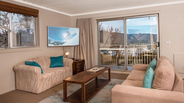 A living room with a sofa, a coffee table, and a large window with a view of a mountainous landscape. - Erulisse (Jindabyne)