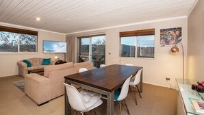 A modern living room with a wooden dining table, beige sofa, and large windows offering views of the outdoors. - Erulisse (Jindabyne)