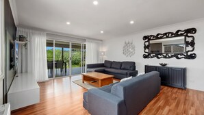 Un salon moderne avec un canapé gris, une table basse en bois et un meuble noir. Une porte-fenêtre coulissante donne accès à un balcon d’où l’on peut admirer la verdure.