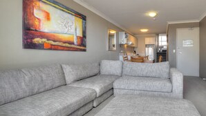 Ein Wohnzimmer mit grauem Ecksofa, einem großen abstrakten Gemälde an der Wand und Blick in den Küchenbereich.
