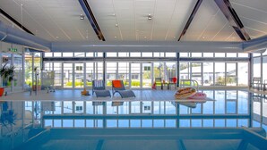 Une grande piscine intérieure avec des parois en verre transparent, de nombreux fauteuils et une vue sur l’extérieur.