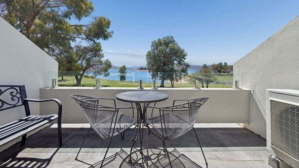 Un balcon avec une table et des chaises en métal, un banc et une vue sur un lac et des arbres.