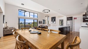 A spacious dining area with a wooden table and chairs, a kitchen with modern appliances, and large windows offering a view of the outdoors.