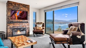 A living room with a fireplace, a large window with a view of a lake and mountains, a leather sofa, and a wooden coffee table.