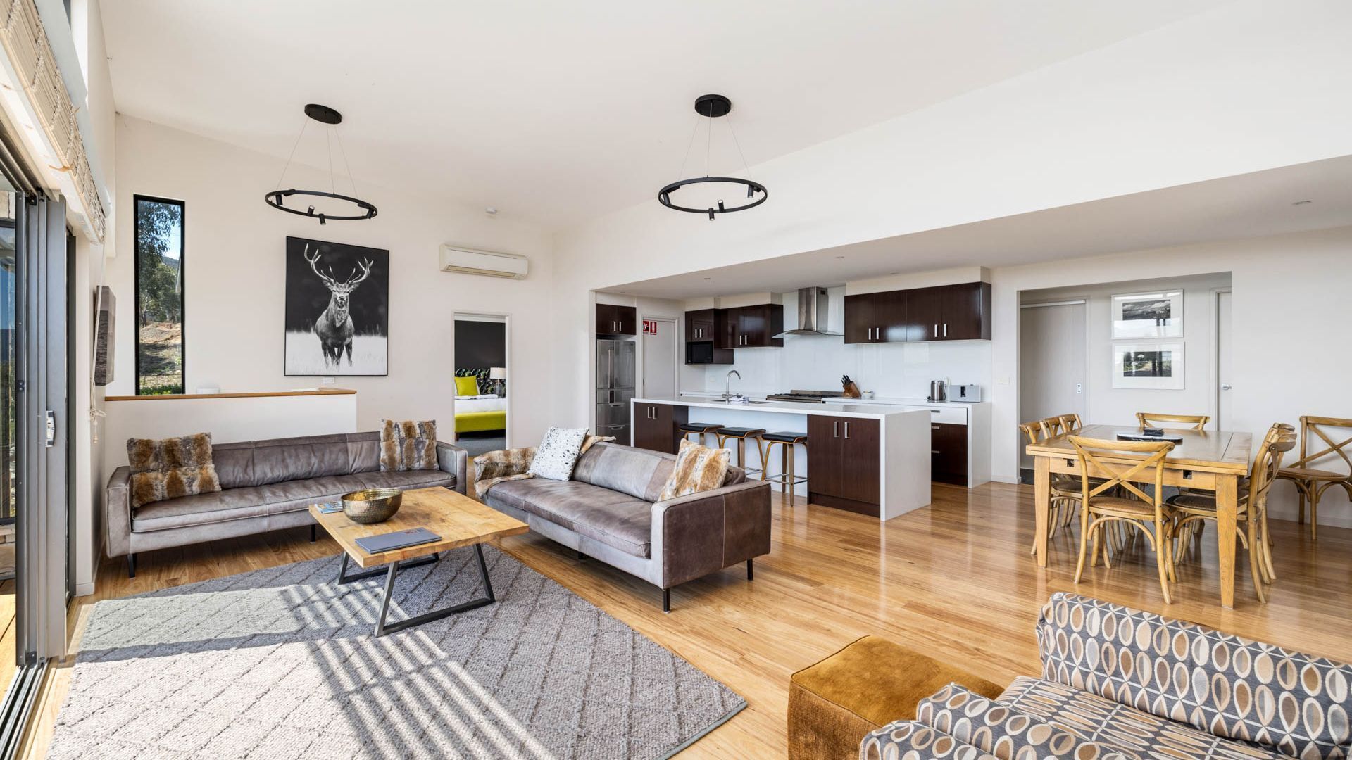 A spacious living area with a sofa, coffee table, and patterned ottoman. A dining area with a wooden table and chairs is visible. The kitchen area includes dark cabinets and a white countertop.