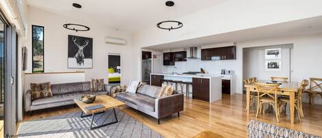 A spacious living area with a sofa, coffee table, and patterned ottoman. A dining area with a wooden table and chairs is visible. The kitchen area includes dark cabinets and a white countertop.